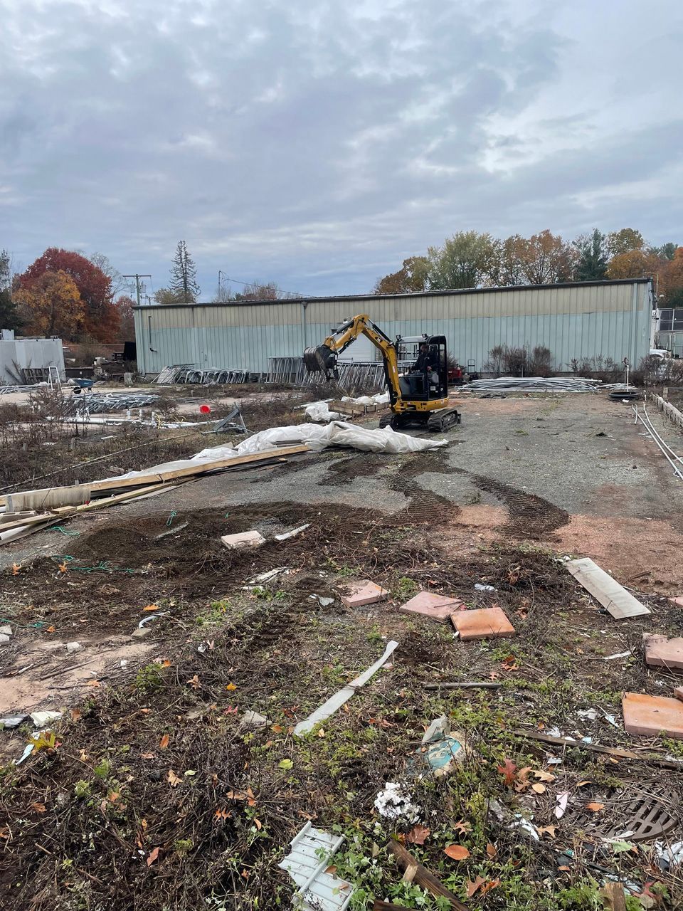 Excavator working in a debris-strewn vacant lot beside a long metal building under a cloudy sky