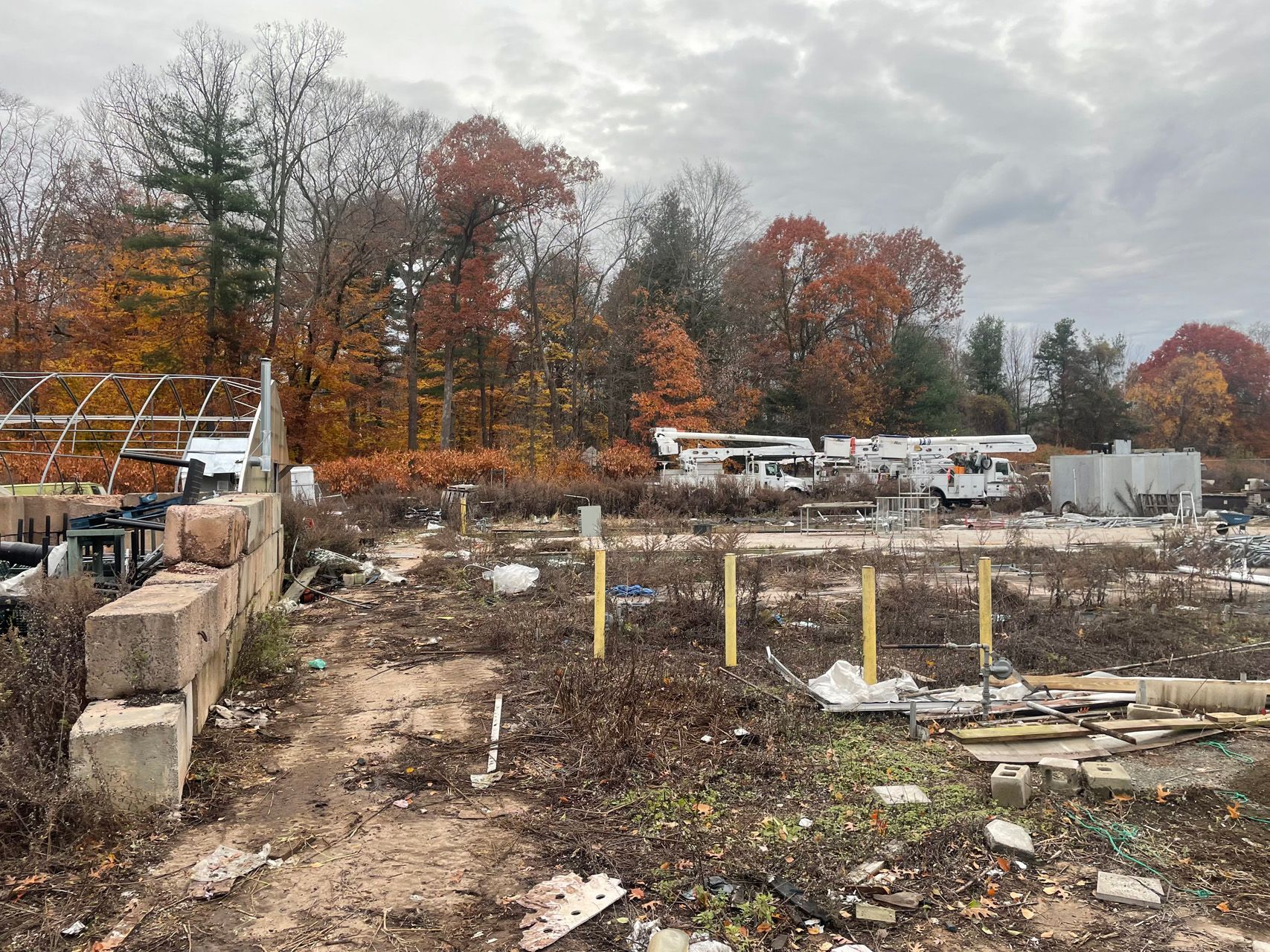 Autumn construction site with dirt lots, concrete blocks, yellow posts, and RVs near trees under cloudy sky