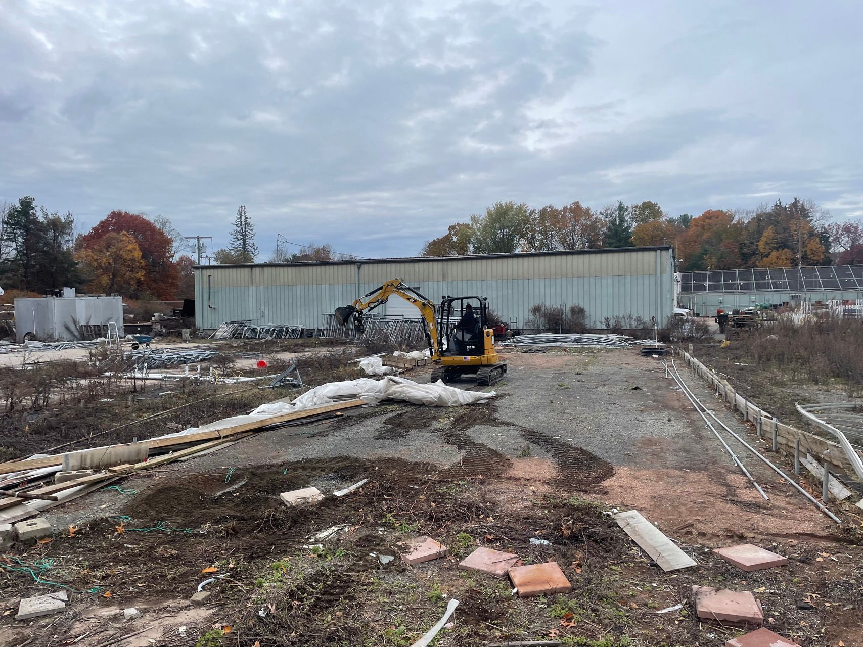 Small excavator on a muddy demolition site with a long white building and cloudy sky in the background