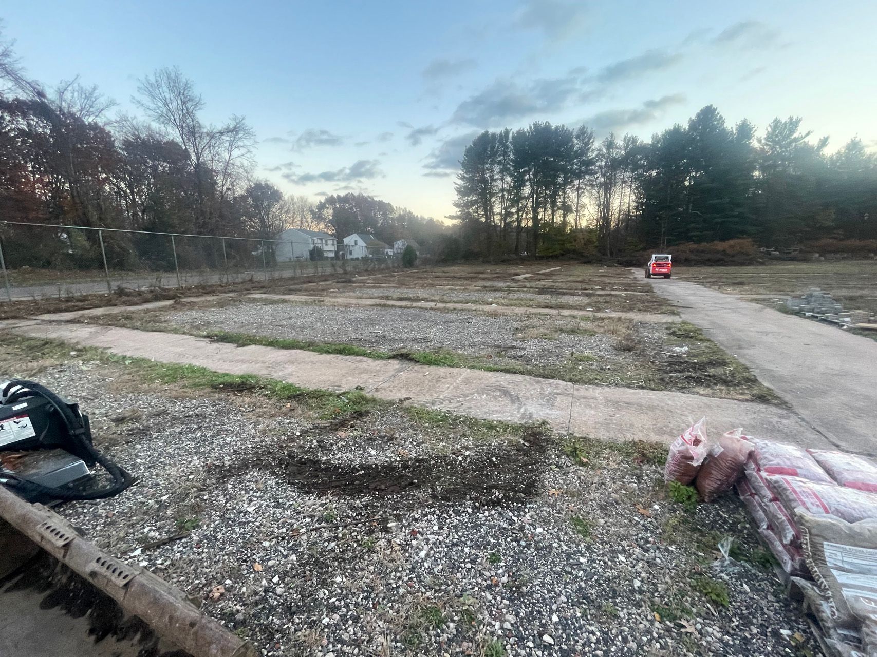 Empty gravel lot with intersecting dirt paths, trees at sunset, and construction debris in the foreground