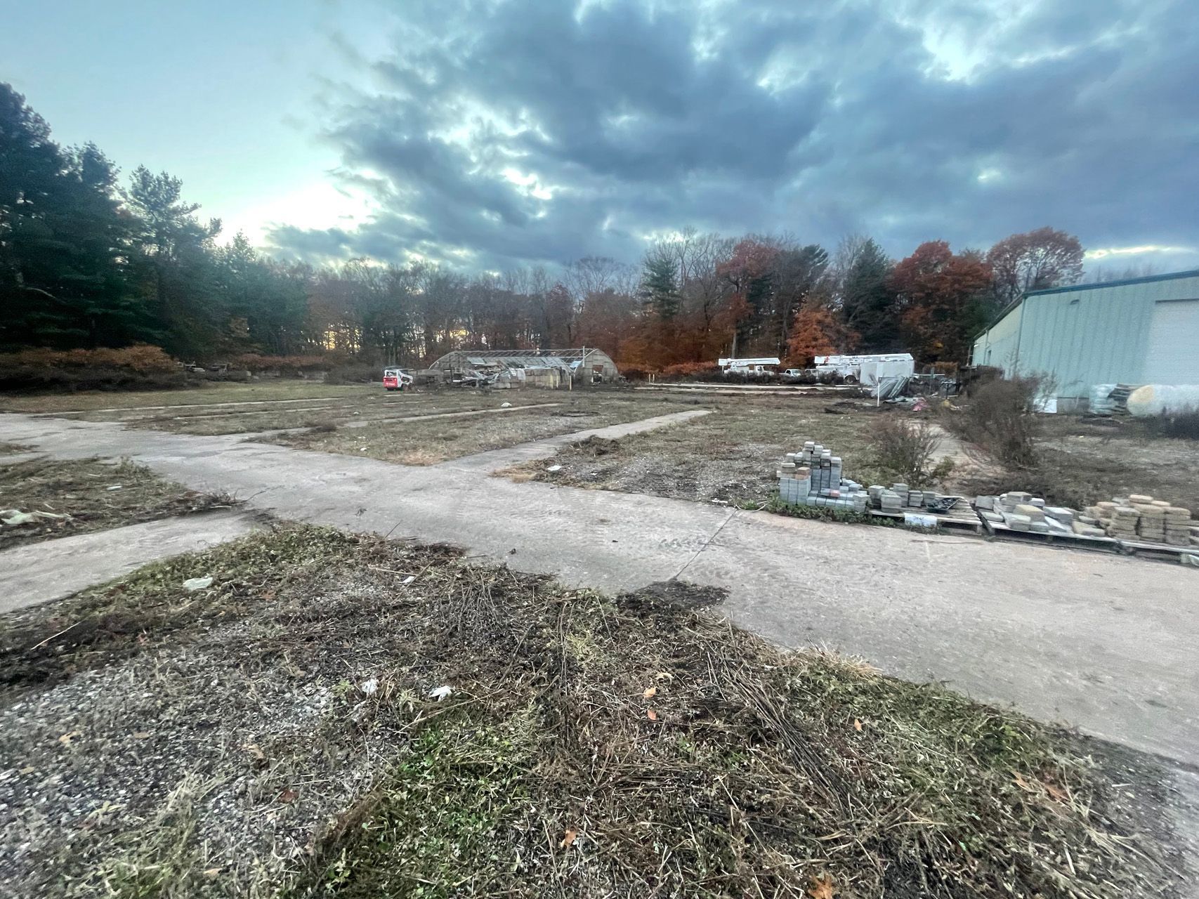 Dirt lot with a gravel path, scattered debris, trees, and a cloudy sky at dusk