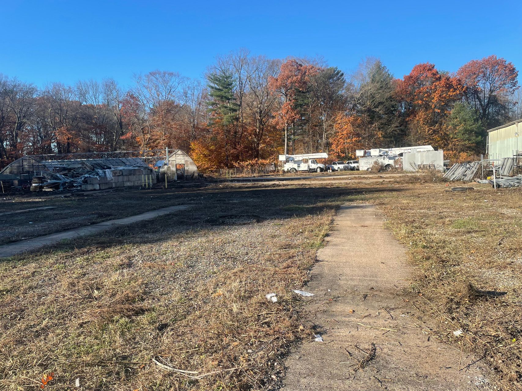 Empty gravel lot with a dirt path, leafless autumn trees, and parked vehicles under a clear blue sky