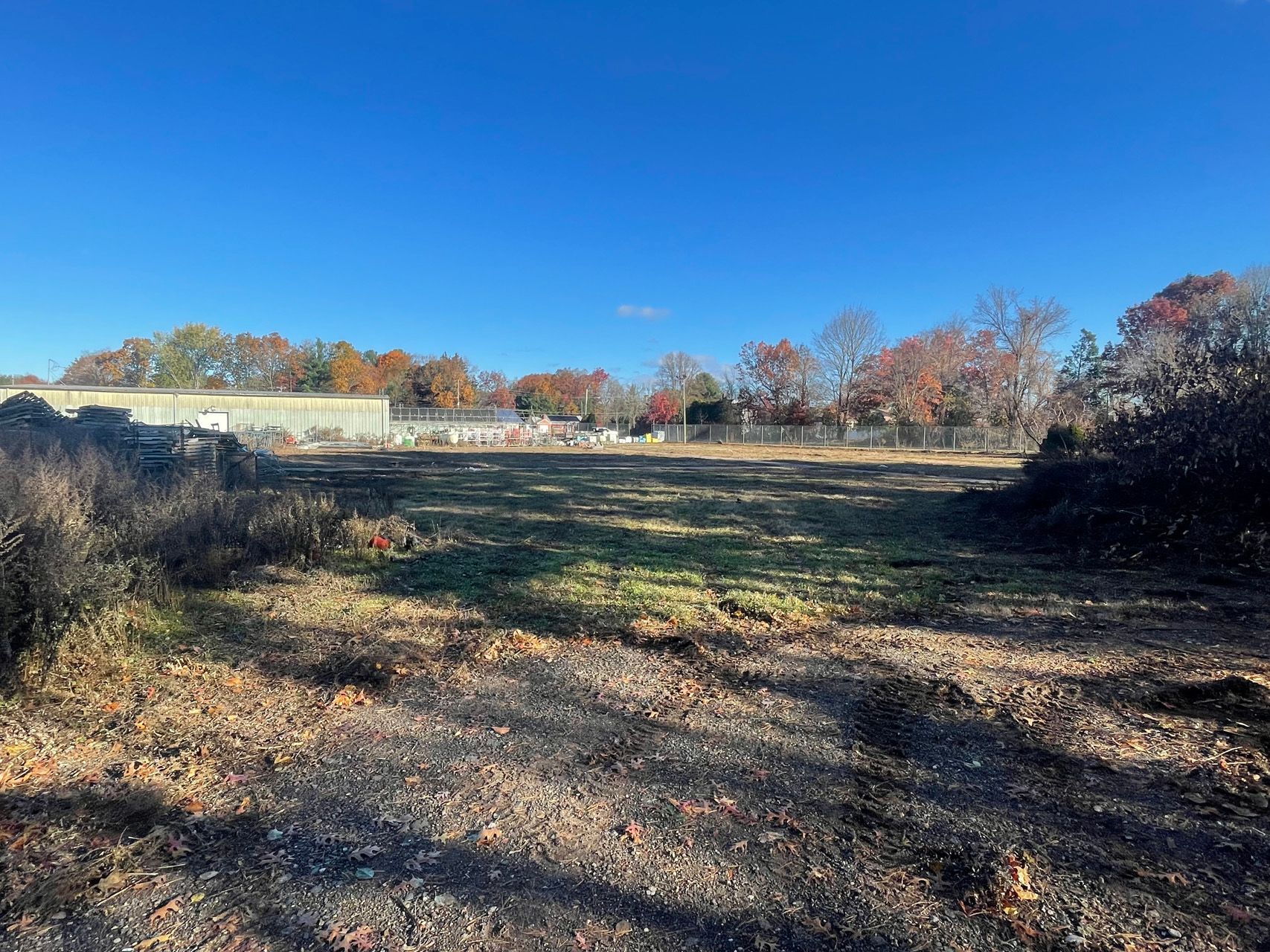 Open field with gravel and brush under a clear blue sky, with trees and a building in the distance