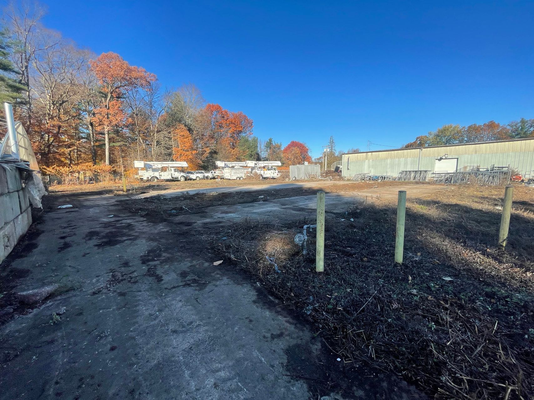 Empty paved lot with black gravel, wooden posts, and autumn trees under a bright blue sky