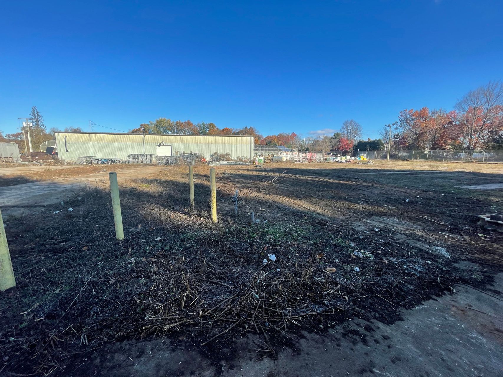 Charred vacant lot with wooden posts under a clear blue sky, with buildings and trees in the background