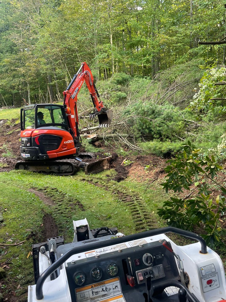 Orange excavator clearing brush on a steep wooded hillside, with another machine in the foreground.
