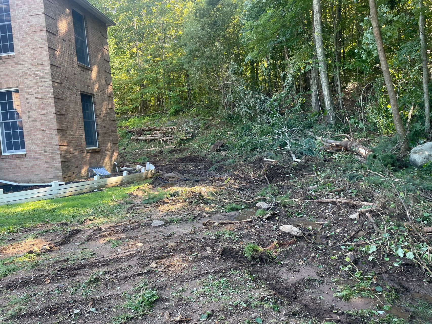 Cleared dirt slope beside a brick house, with trees and forested yard in the background