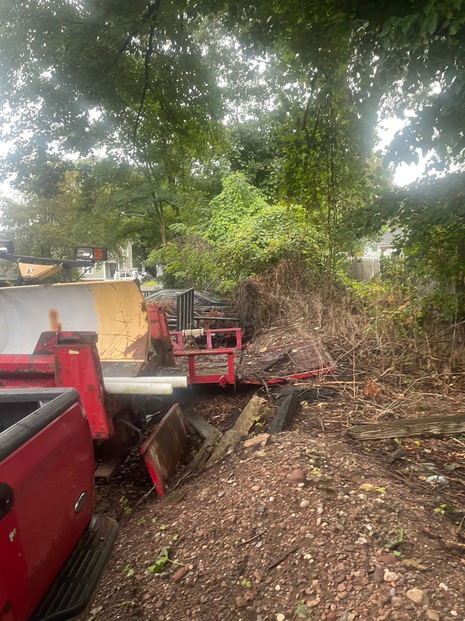Red dump truck loading debris from a wooded roadside pile beside a wall
