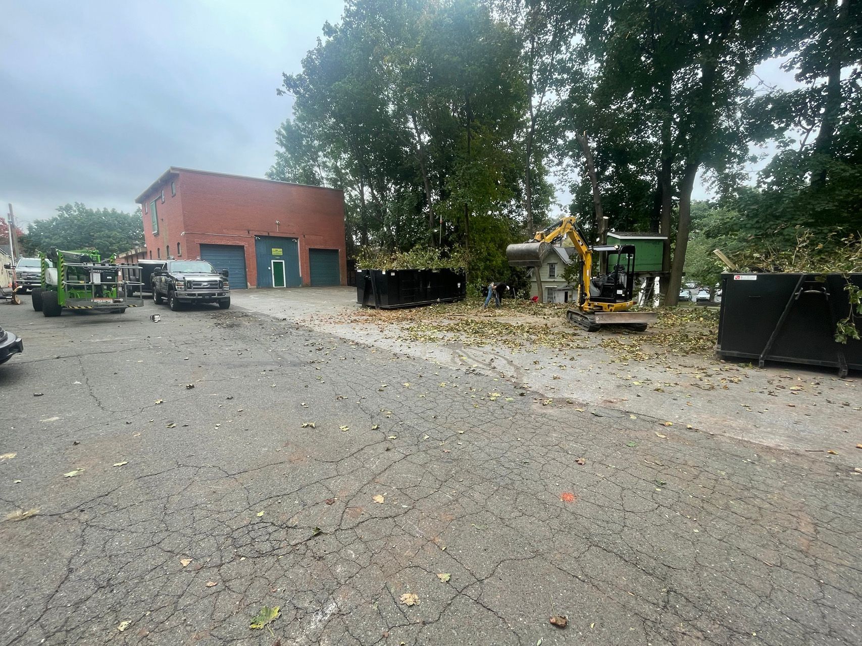 Construction site with excavator, trucks, and a red building on a wet gravel lot