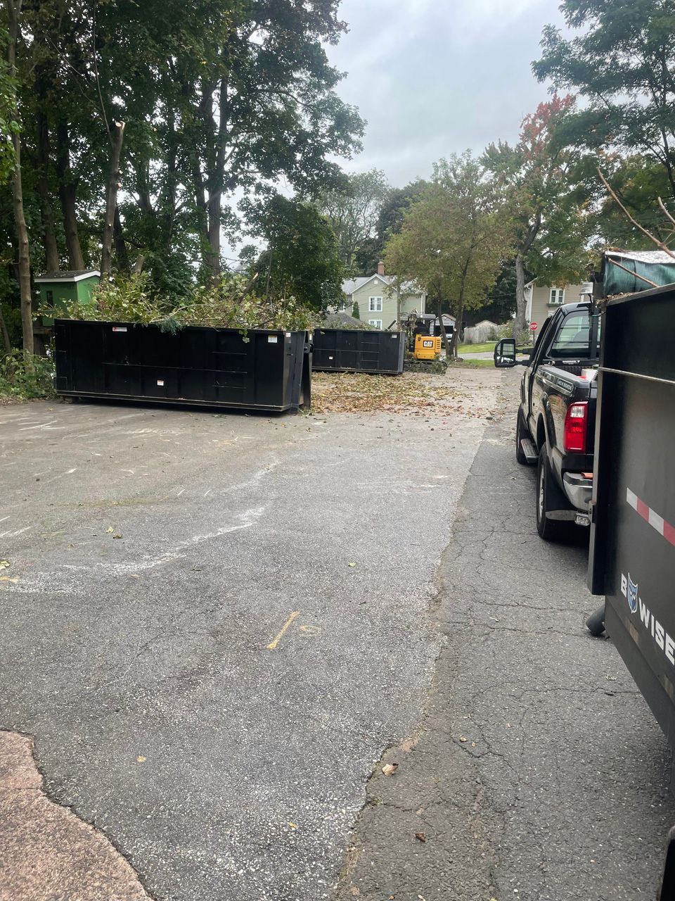Outdoor parking lot with black dumpsters, trucks, and scattered leaves under trees