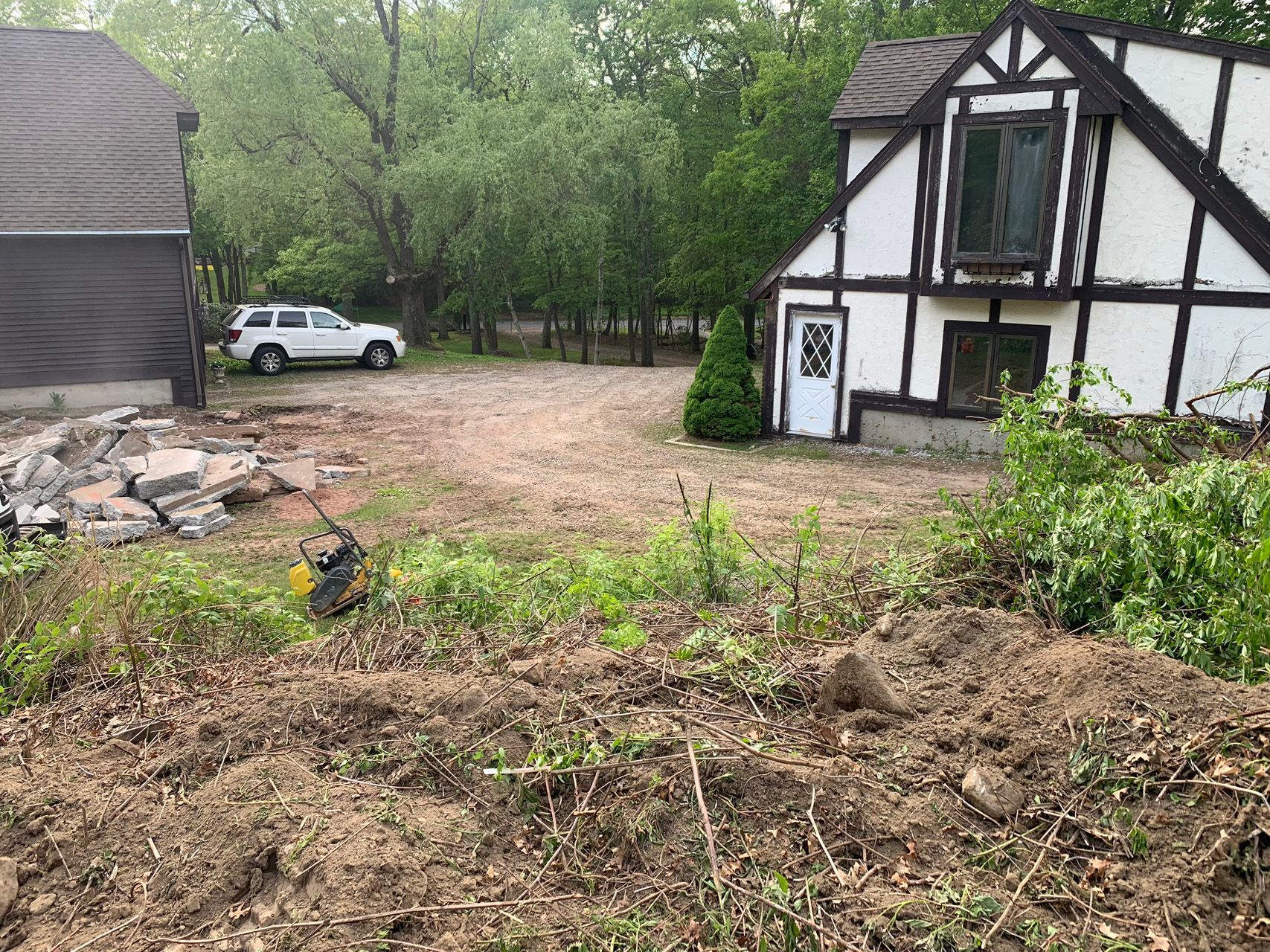 Rural yard with a white house, white SUV, and a pile of debris near a muddy garden bed