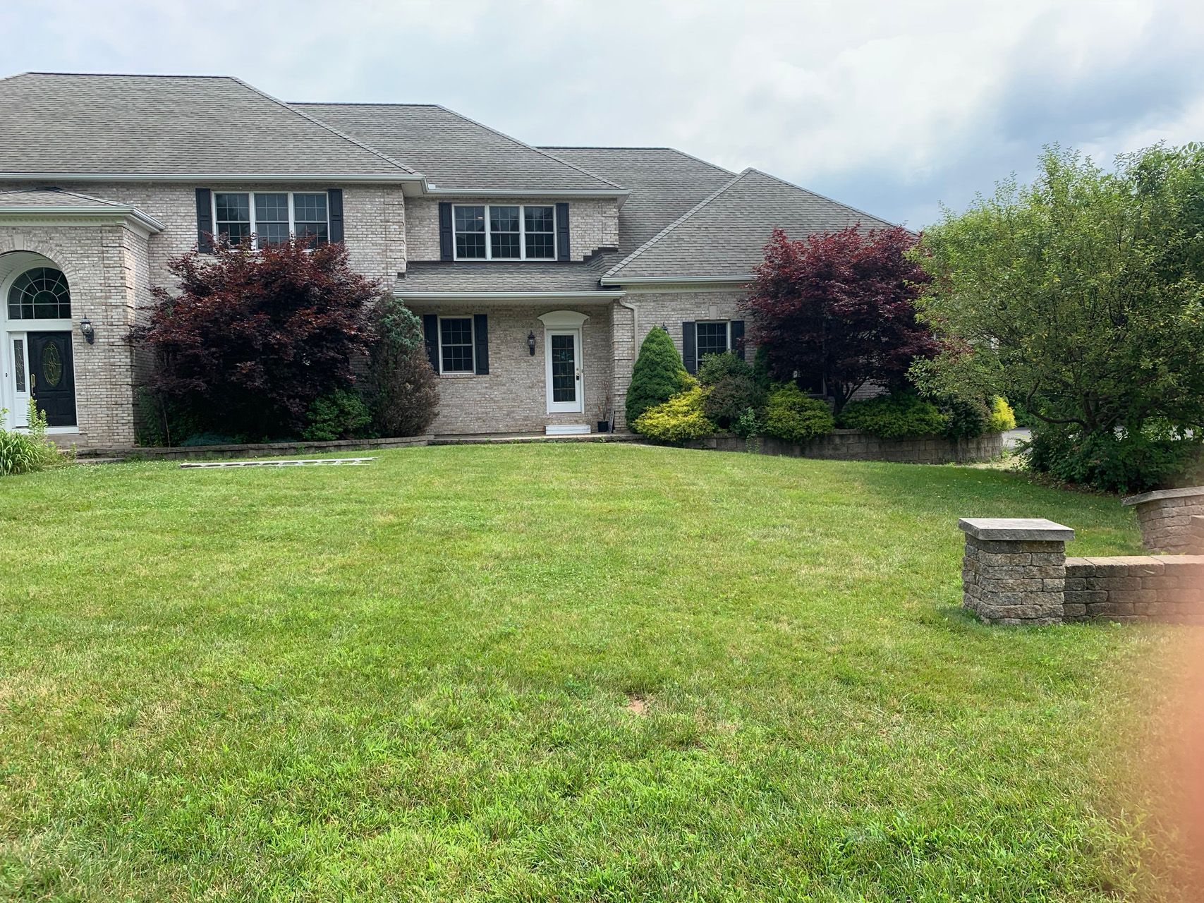 Suburban house with a large green lawn, shrubs, and a stone mailbox pillar under cloudy sky