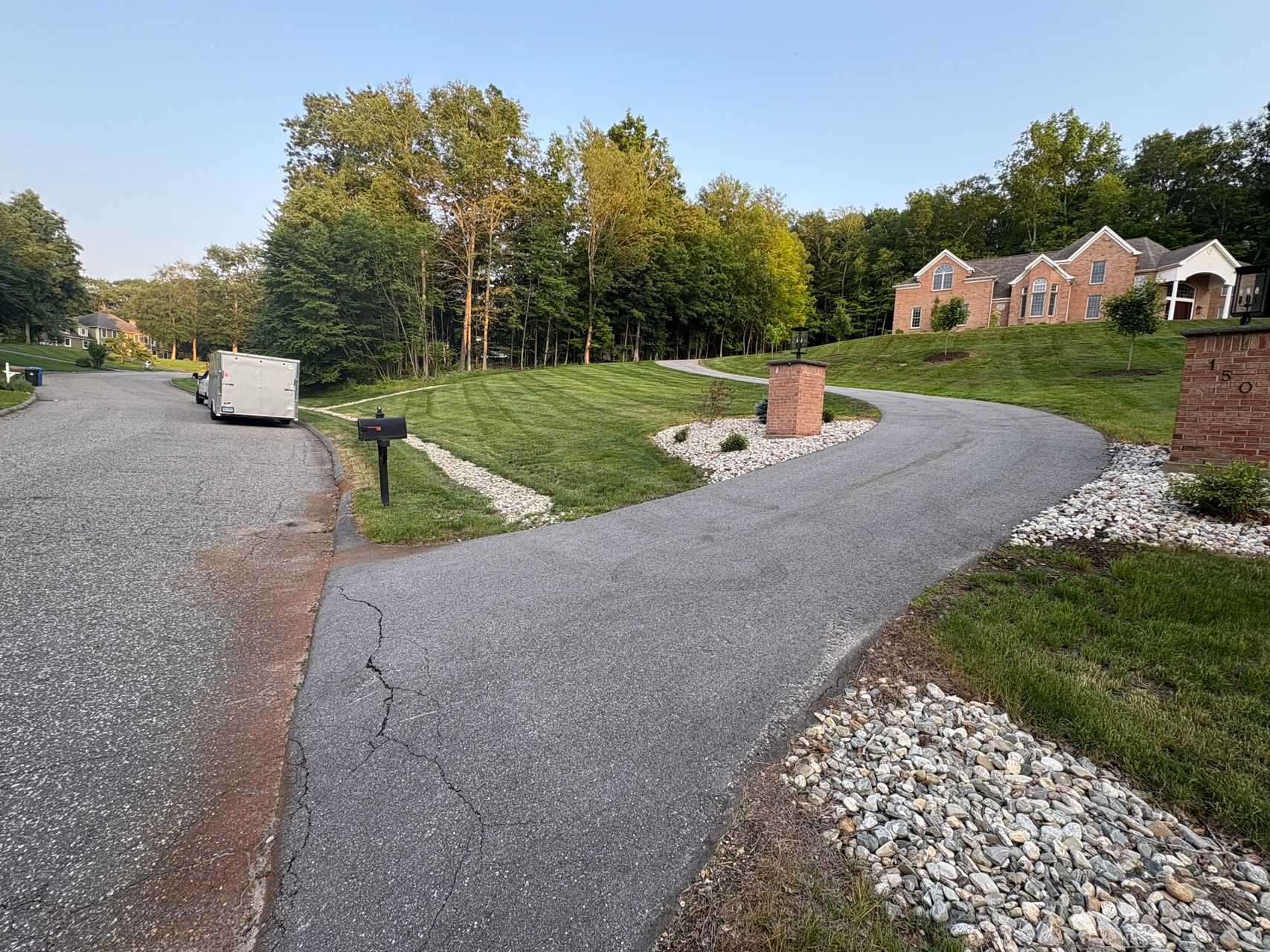 Gravel driveway curves past landscaped yard and brick pillars toward a house in a wooded neighborhood