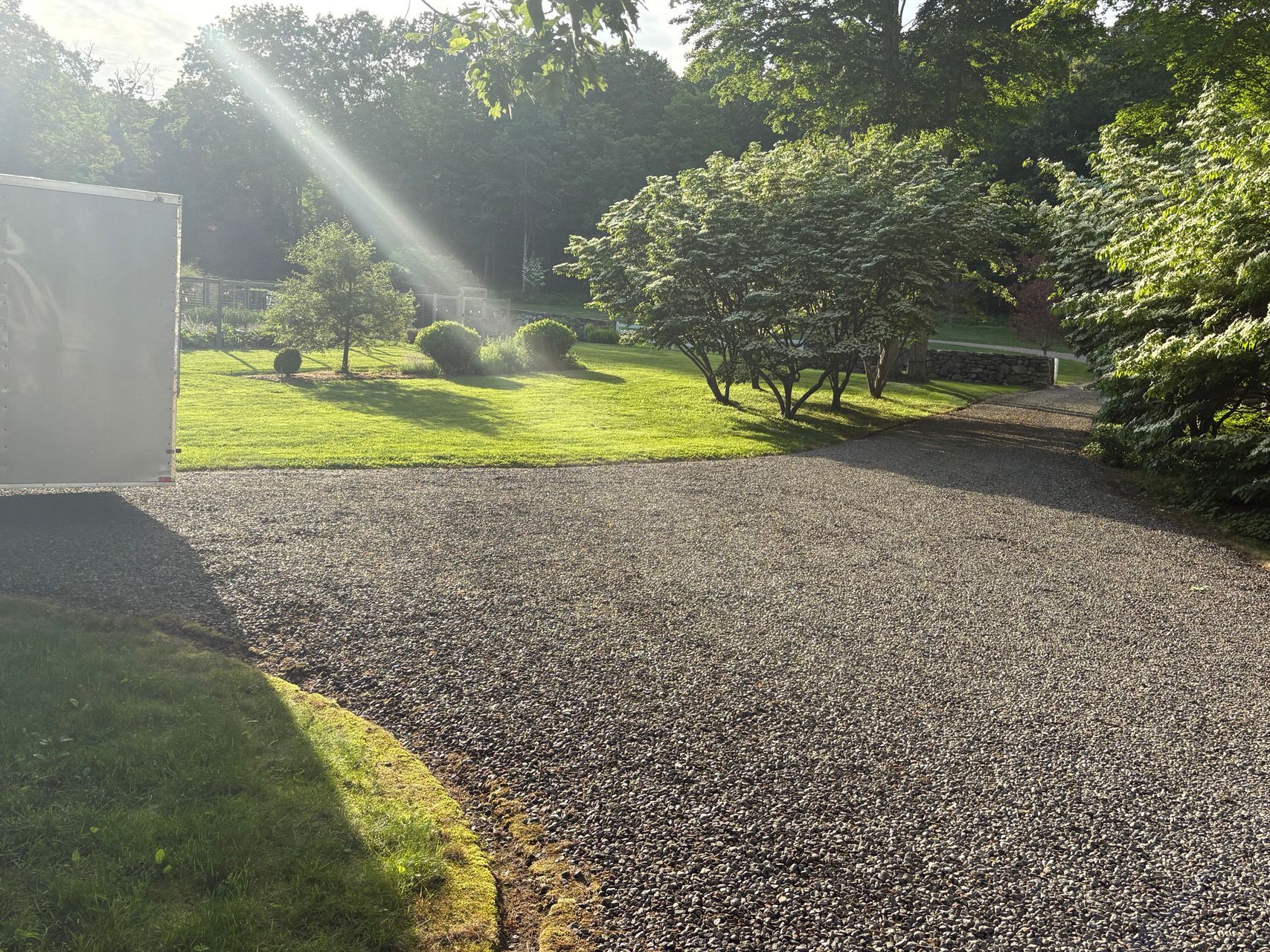 Sunlit gravel driveway beside green shrubs and trees, with a white wall on the left