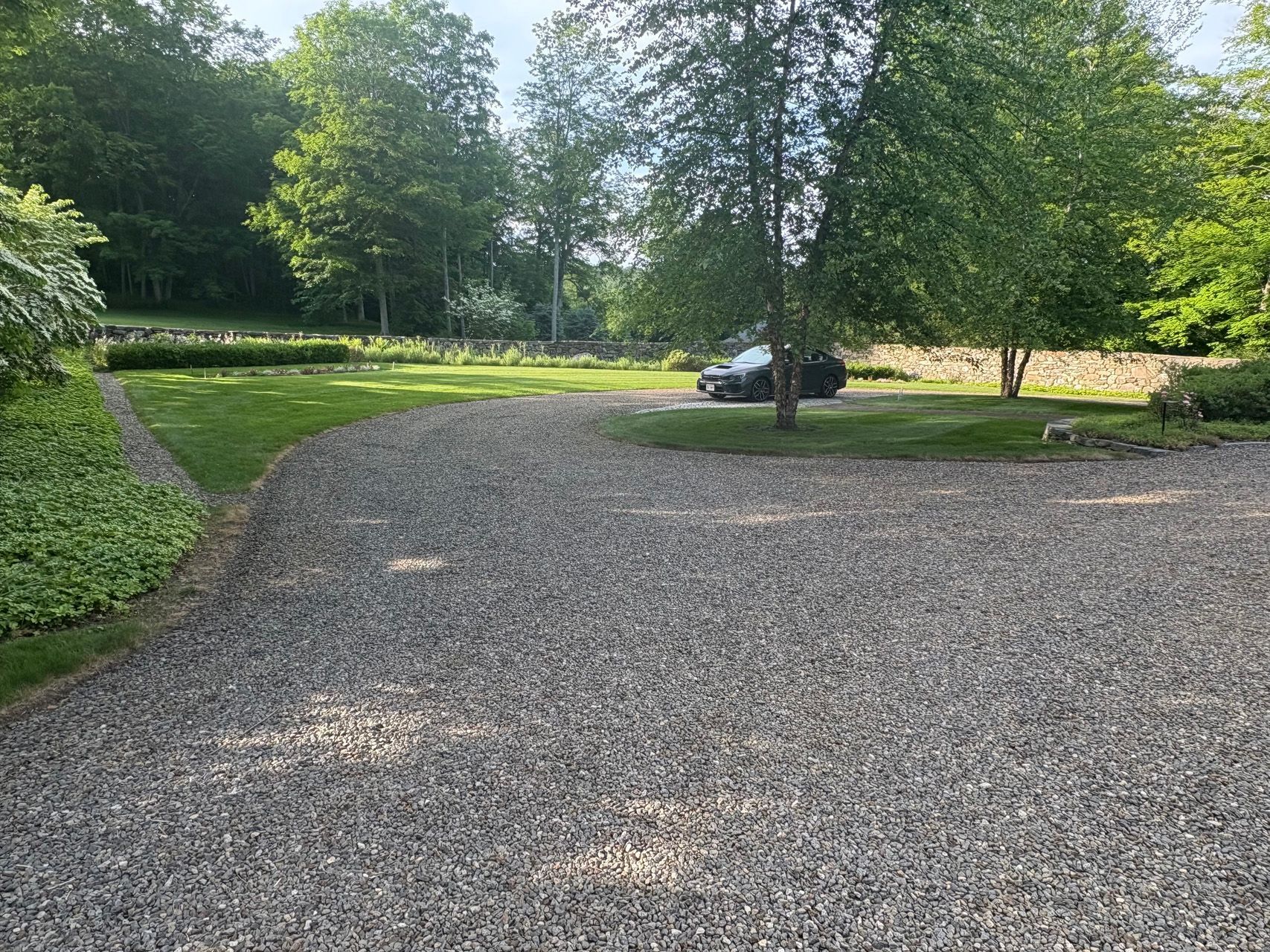 Gravel driveway curving through a green wooded yard with a parked car in the distance.