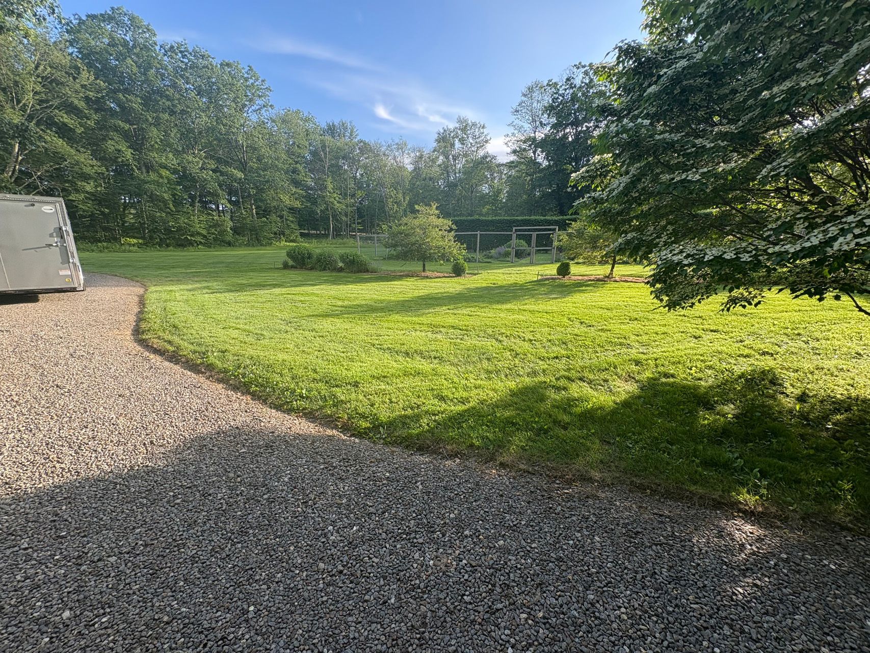 Gravel driveway beside a sunny grassy yard with trees and a small fenced area in the distance