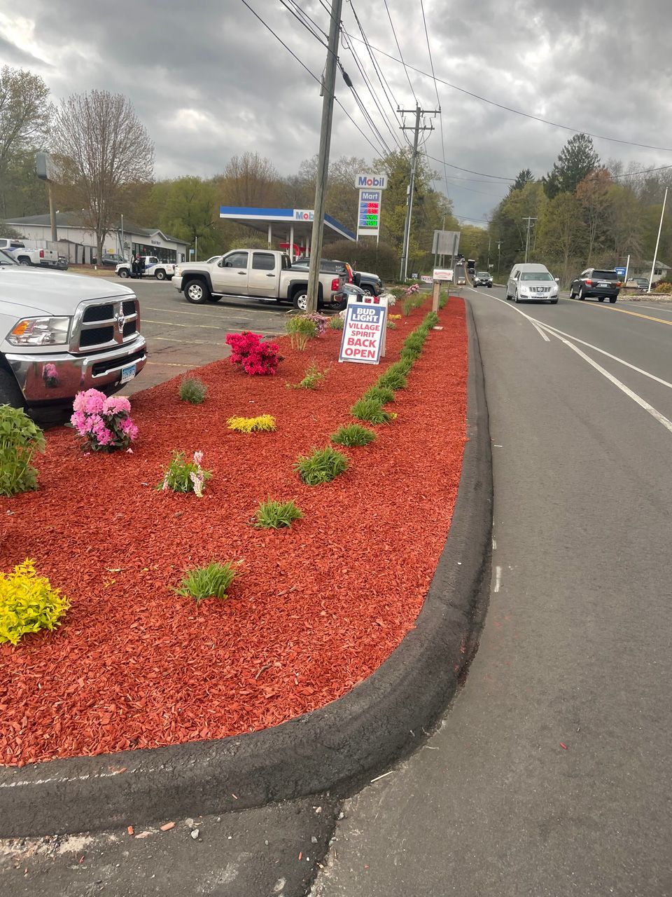 Parking lot with red mulch flower bed, pink decorations, and road beside a business sign on an overcast day
