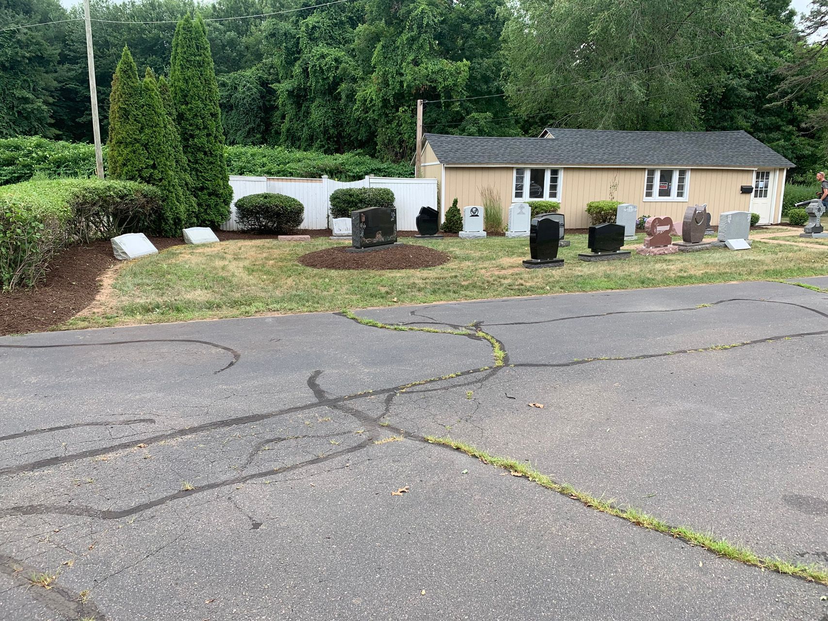 Cracked asphalt driveway leading to a small tan house with garage, shrubs, and trees in a quiet neighborhood