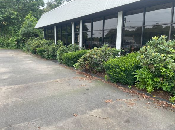 A small roadside building with a white roof, dark pillars, and green bushes along a paved road.