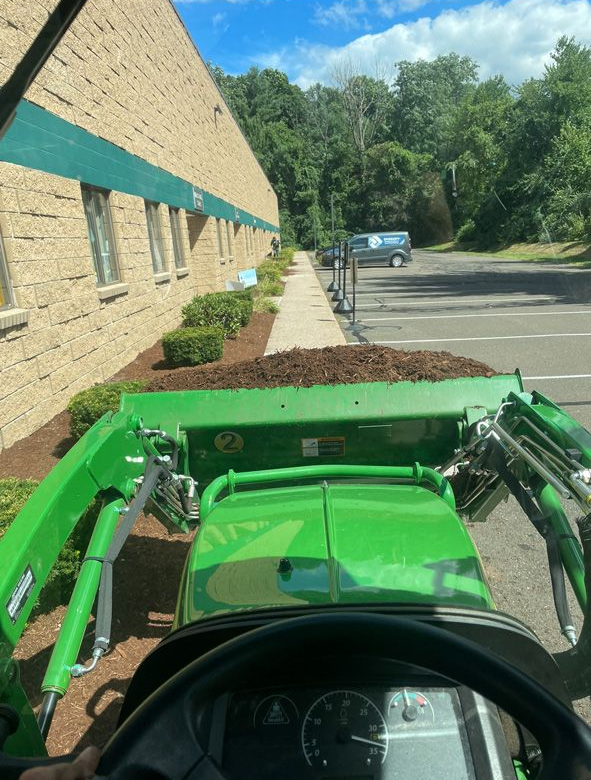 Green tractor loader driving beside a tan brick building on a sunny street with trees ahead