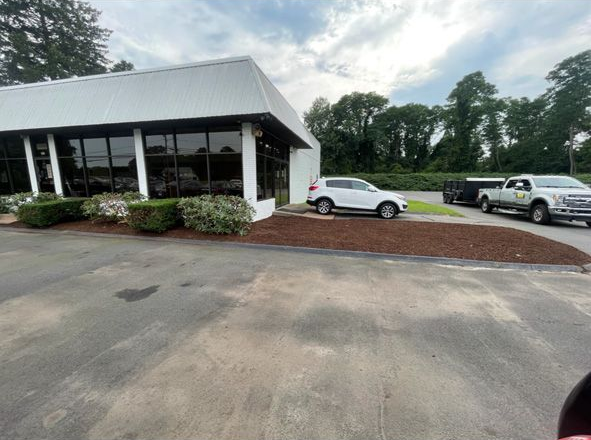 Small white building with a covered porch, parked car, and red gravel lot under a cloudy sky