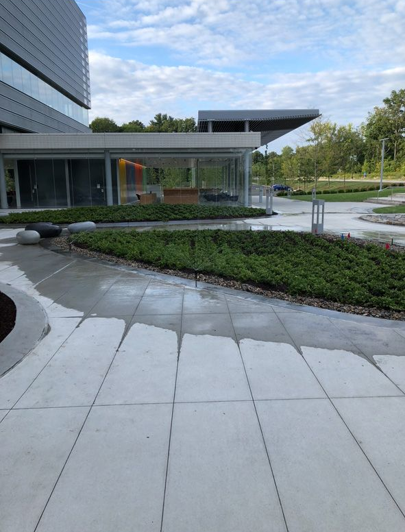 Modern building entrance with curved stone walkway, landscaped greenery, and blue sky.