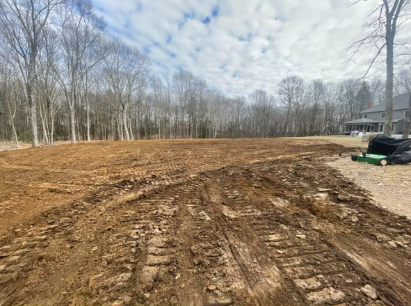 Dirt lot with tire tracks beside leafless trees under a cloudy sky, with a parked vehicle at right.