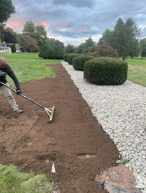Worker spreading soil beside a white gravel path in a landscaped yard under cloudy skies