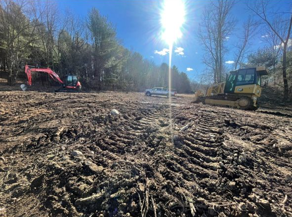 Sunlit dirt lot with wood chips, trees, a pink excavator, and a small building in the background