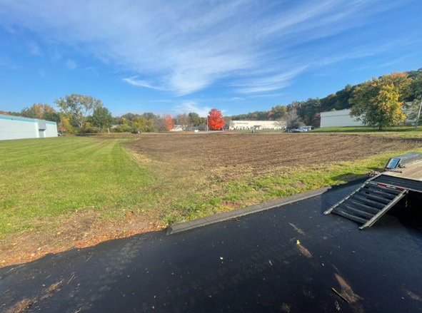 Open field with a red barn under a blue sky, seen beside a paved path and grassy hillside.