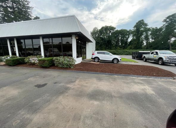 Low white building with a porch and parked car beside a red dirt lot under cloudy skies