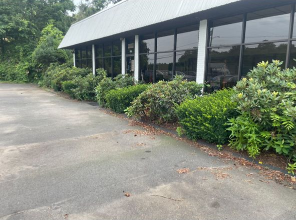 Covered roadside walkway beside a building, with bushes and a paved drive in the foreground.