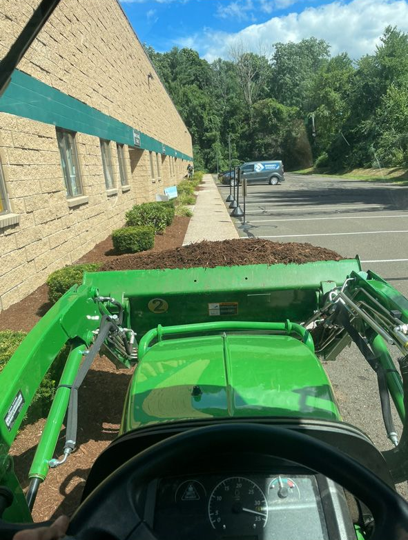 View from a green tractor driving beside a brick building and trees on a sunny day