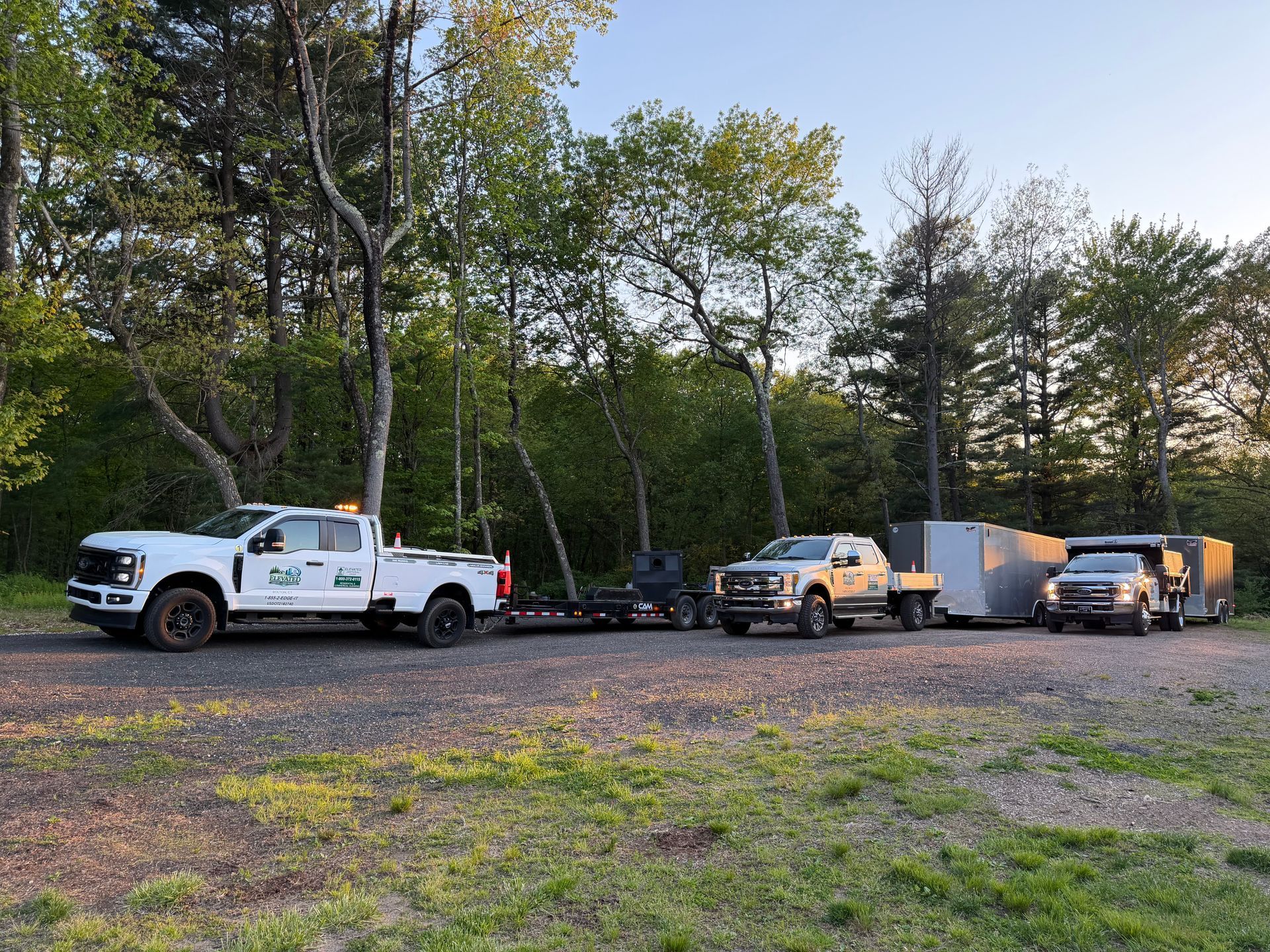 White utility truck, pickup, and enclosed trailer parked on a gravel lot beside trees.