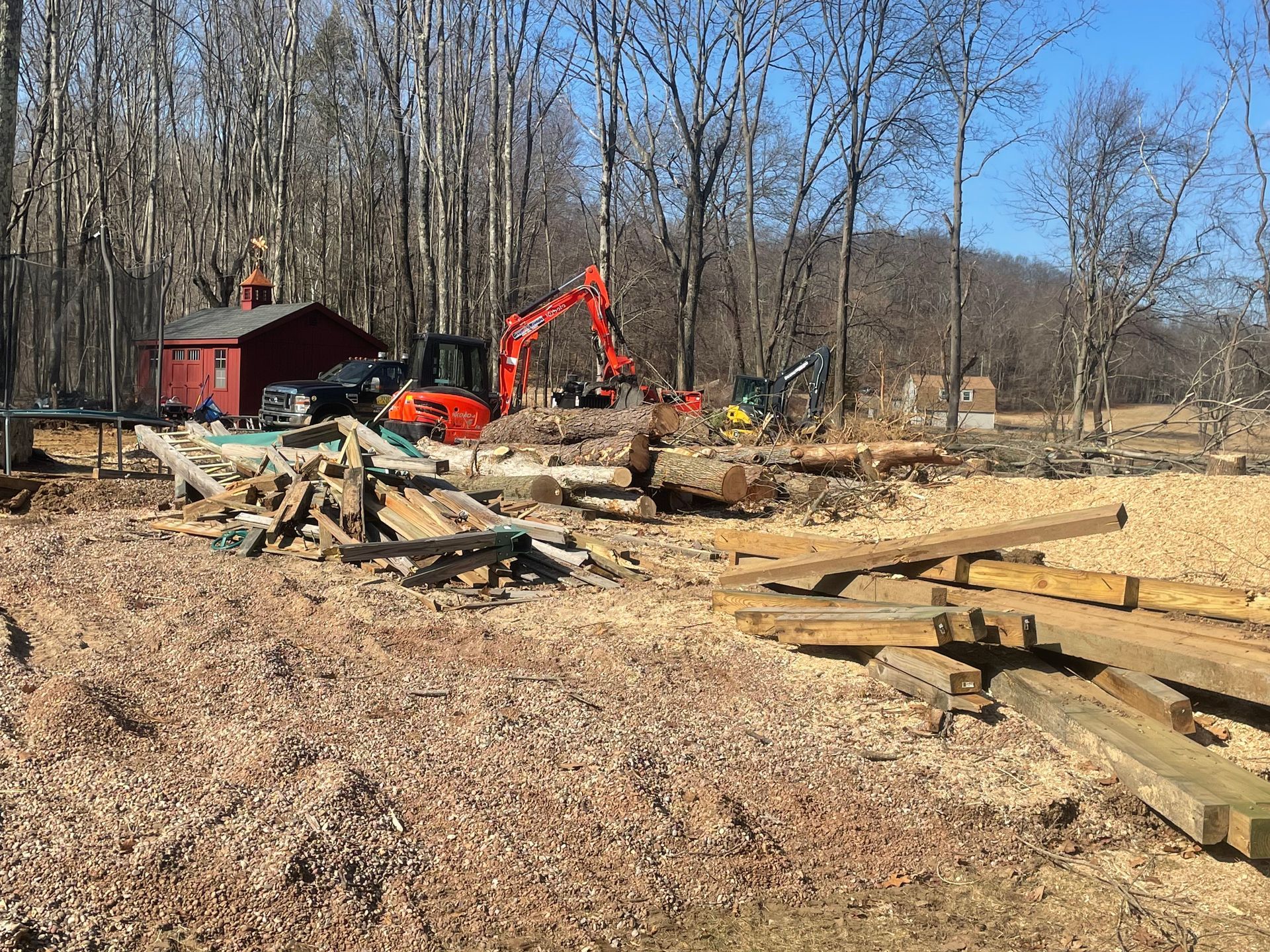 Orange excavator beside red shed in a cleared wooded lot with piles of lumber and debris