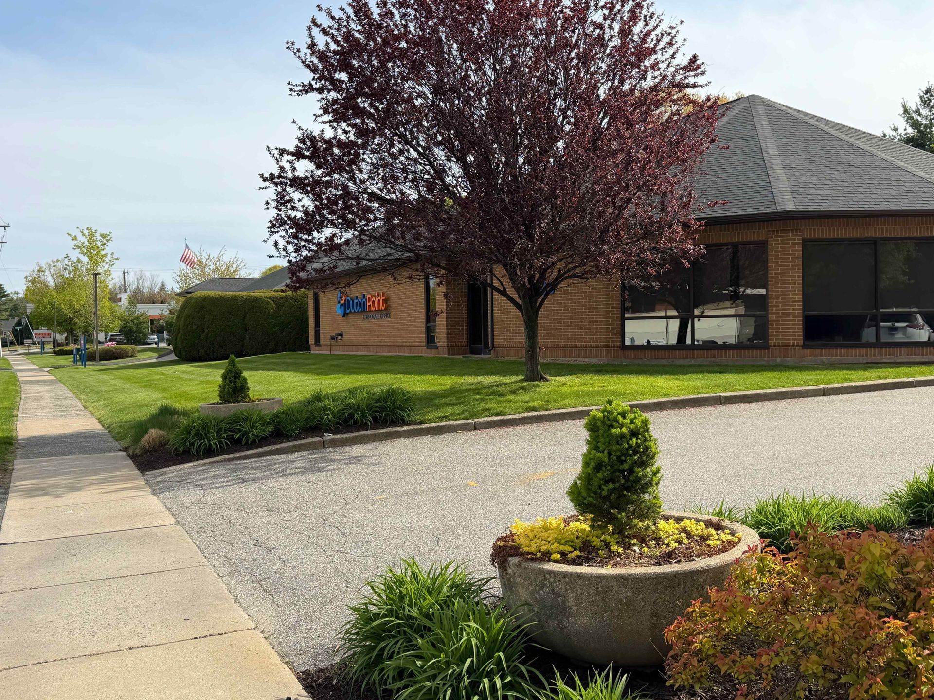 Sidewalk beside a brick building with a dark roof, trees, lawn, and a gravel drive with planters