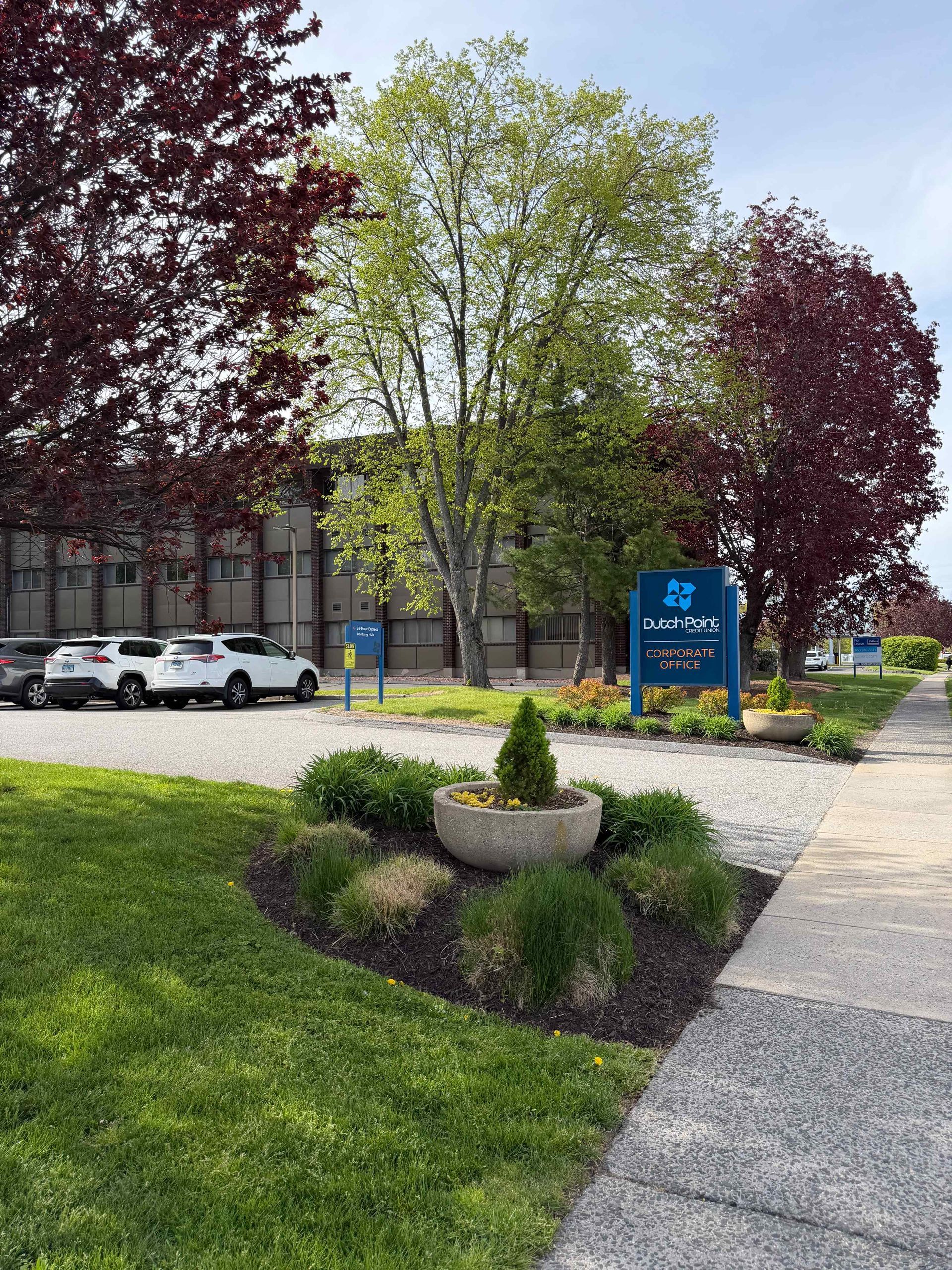 A landscaped sidewalk with trees, grass, flowers, and a blue sign beside a building.