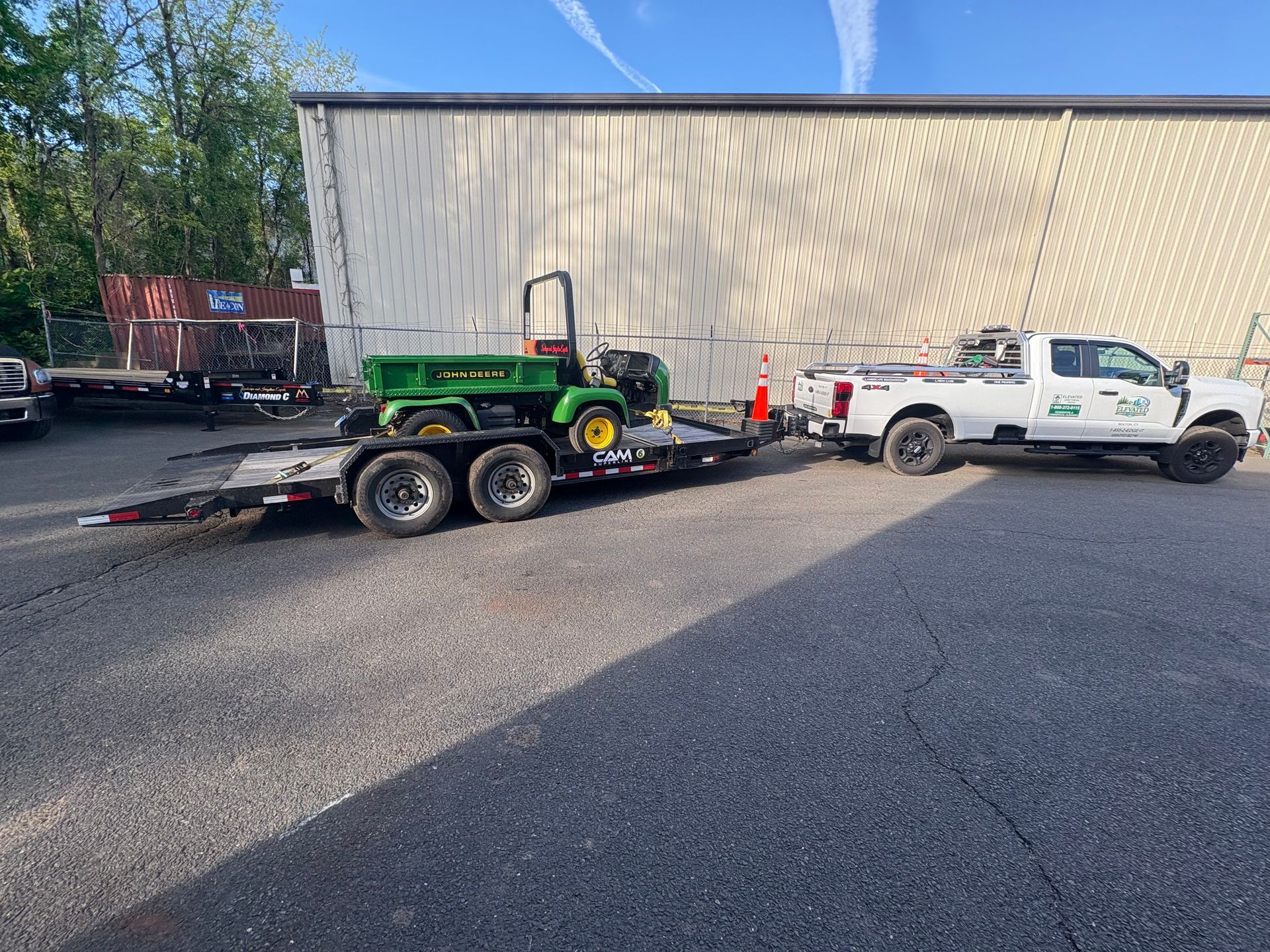 White utility truck towing a green tractor on a flatbed trailer in a parking lot