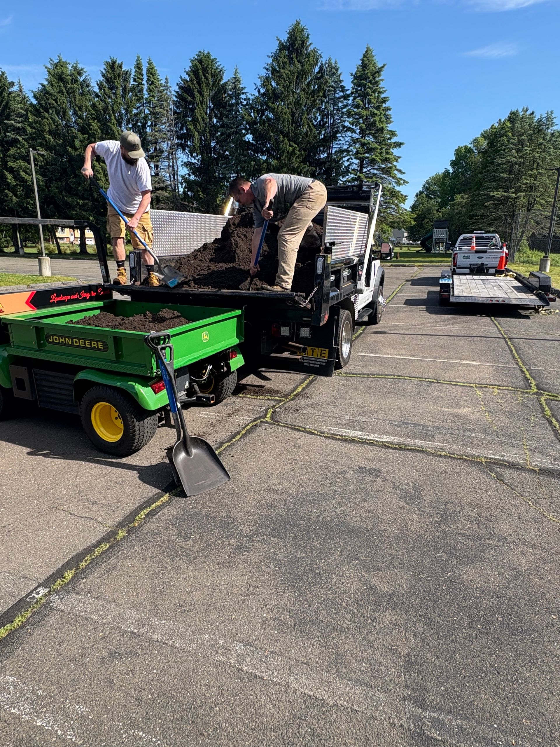 Workers loading gear onto a trailer and truck in a sunny parking lot, with a small green utility vehicle nearby.