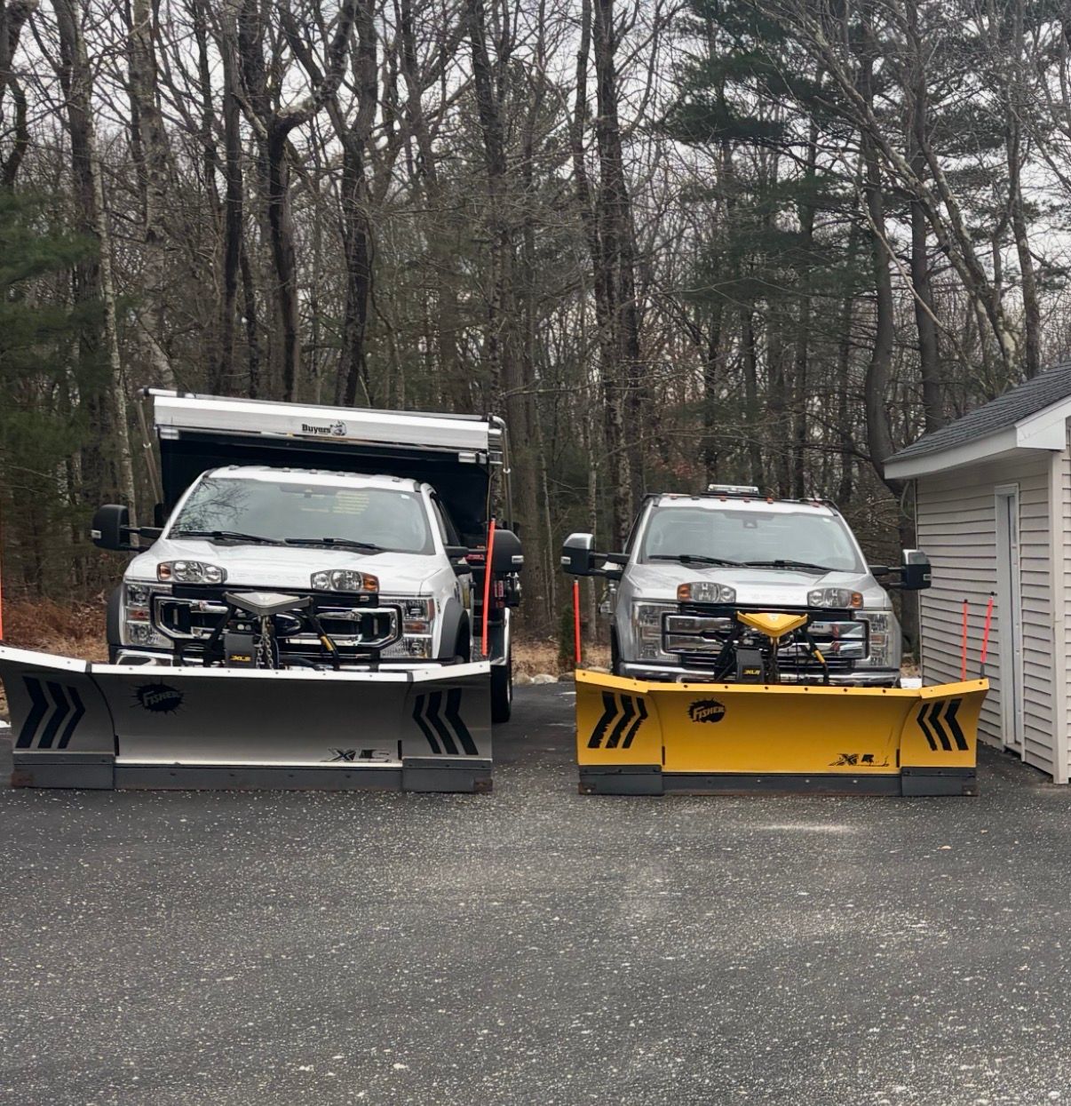 Two pickup trucks with snowplows parked on a paved area, with trees in the background. One plow is silver, the other yellow.