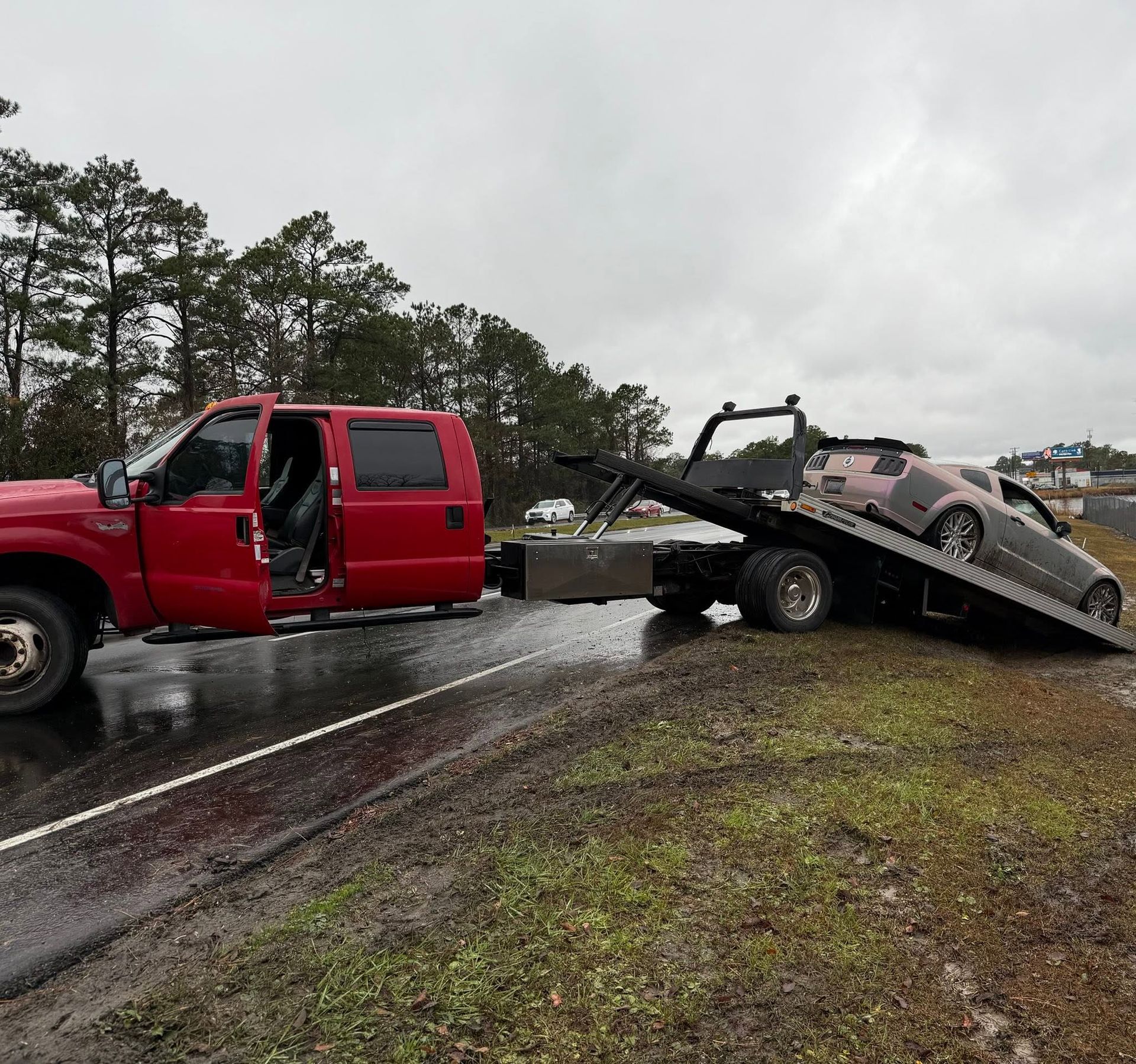 Red tow truck hauling a mangled car on a flatbed beside a wet roadside under gray skies