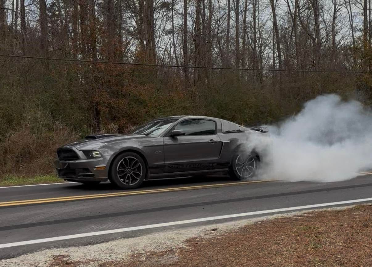 Gray sports car doing a burnout on a wet road, with thick white smoke billowing behind it.