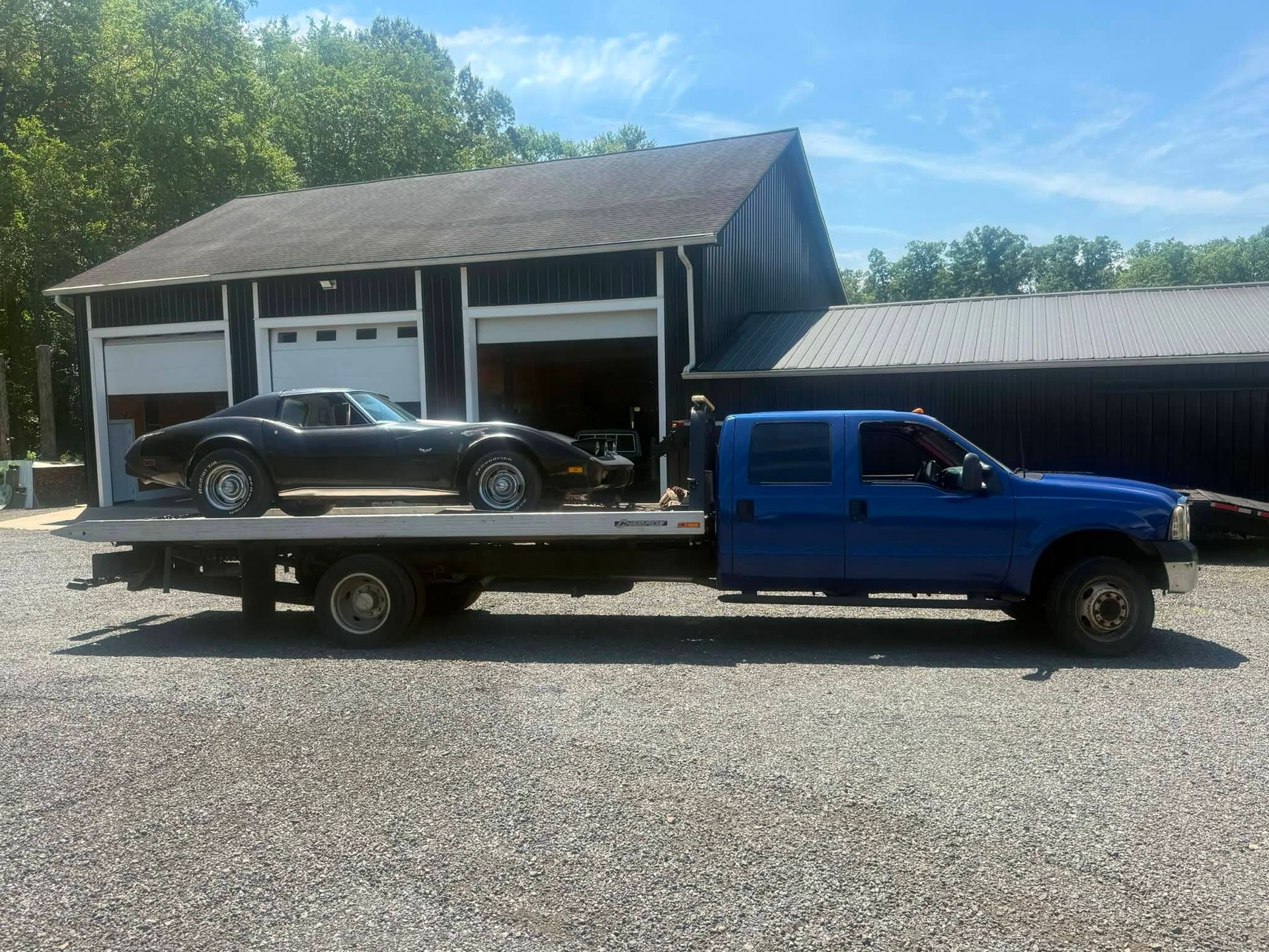 Blue flatbed tow truck hauling a black car in front of a garage.