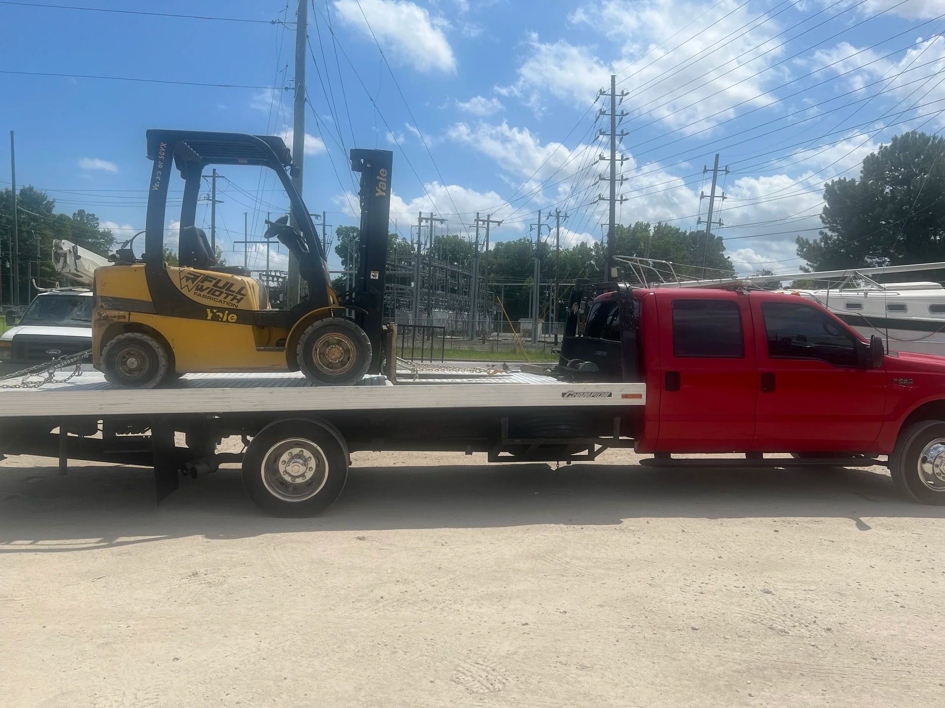 Red flatbed tow truck carrying a yellow forklift on a sunny street.