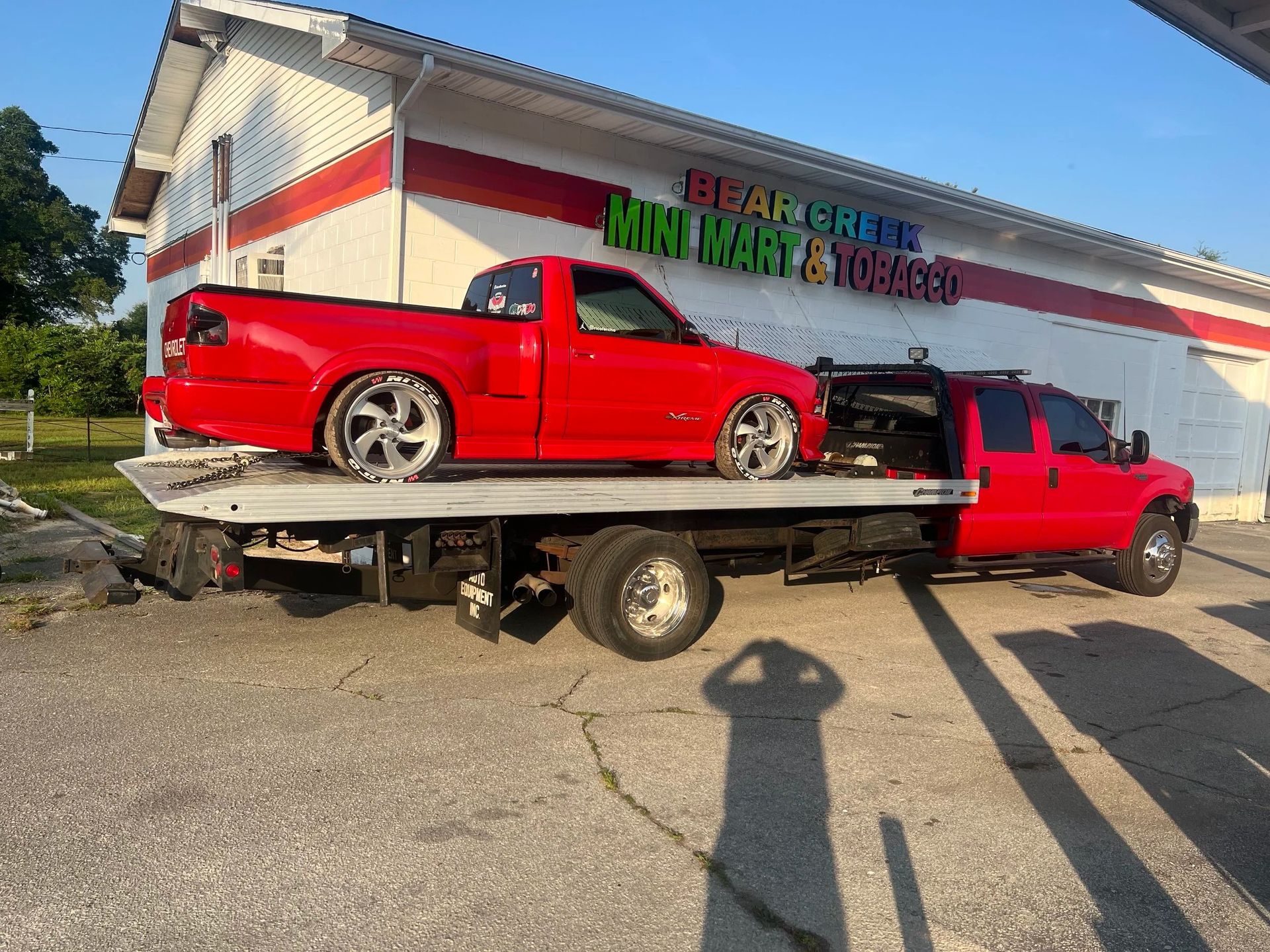 Red car on a tow truck beside a building with a large sign, sunny outdoor scene