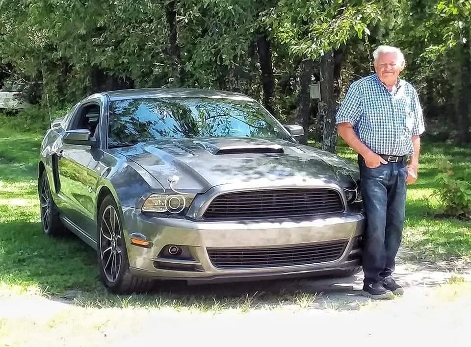 Man standing beside a gray sports car on a tree-lined driveway