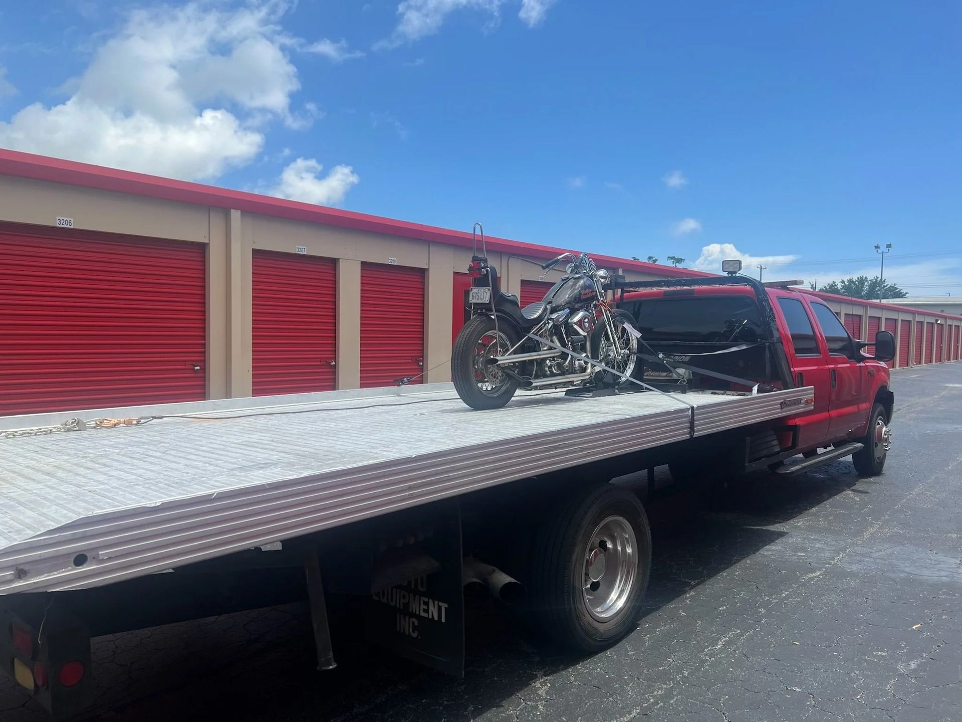 Black flatbed tow truck carrying a red motorcycle in front of red storage units under a blue sky