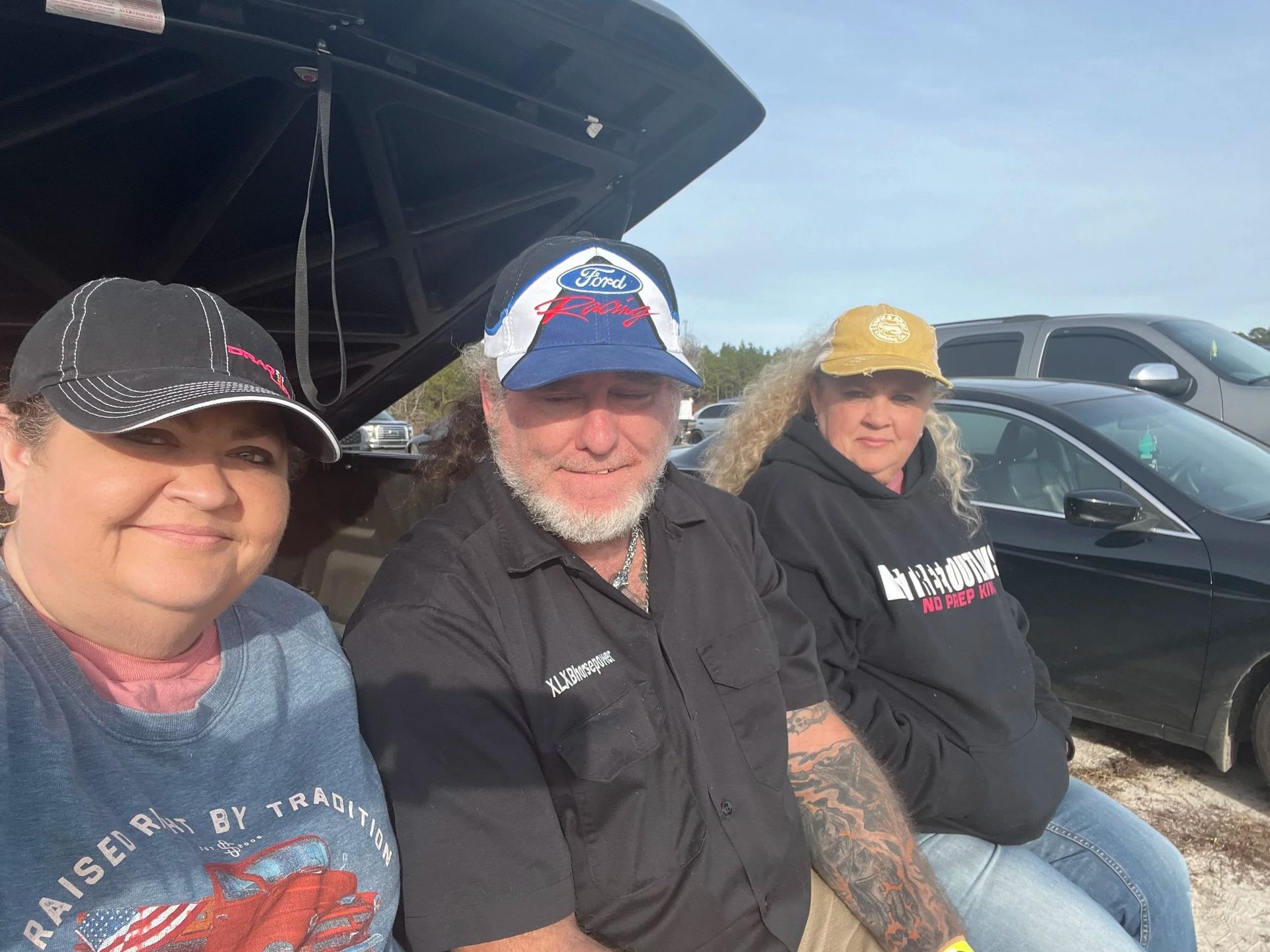 Three people sitting together outdoors beside a car under an open canopy.