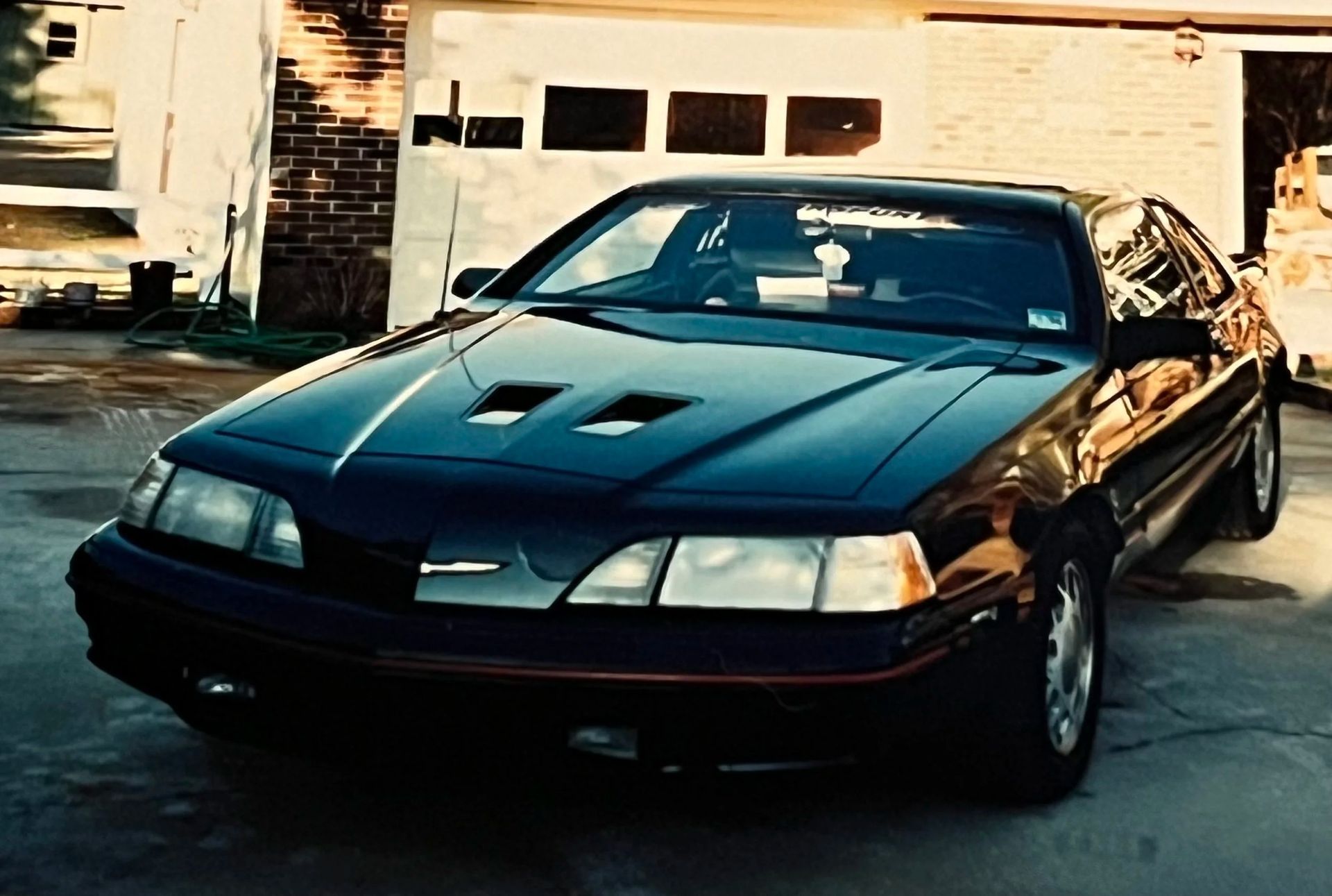 Black sports car parked in a driveway, front view, with house and garage in the background.