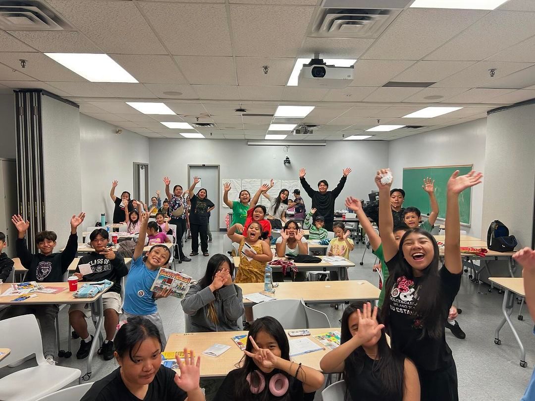 A group of children are sitting at tables in a classroom with their hands in the air.
